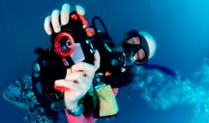 A scuba diver wearing a white helmet and a mask is underwater holding a camera with a red lens. Bubbles rise around them in the blue water, and they appear to be taking a photograph or video.