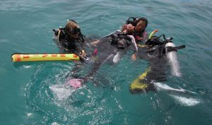 Three scuba divers in the ocean are performing a rescue drill. One diver is holding a floating buoy, while another assists a third person lying face up in the water. Bubbles rise around them, and the water is clear and blue.
